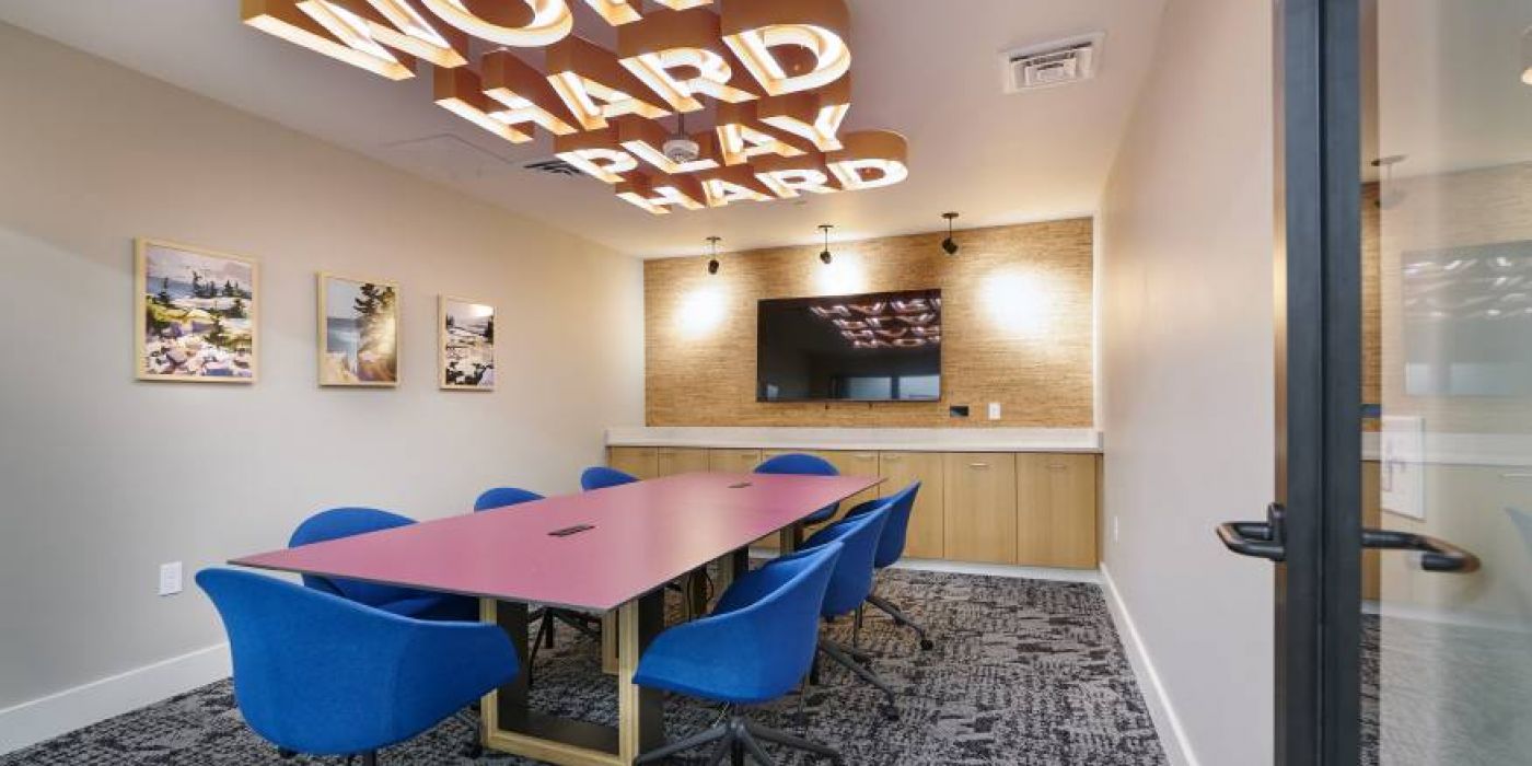 A sleek conference room at VERVE Boise with a vibrant "Work Hard Play Hard" sign, blue chairs, and a wall-mounted TV.