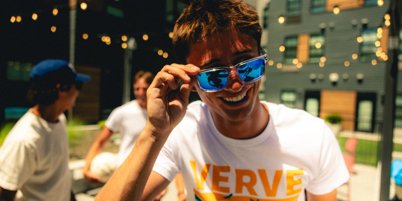 Smiling person in sunglasses and a "VERVE" shirt at an outdoor gathering with friends and string lights.