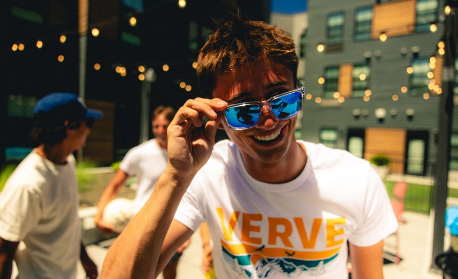 Smiling person in sunglasses and a "VERVE" shirt at an outdoor gathering with friends and string lights.