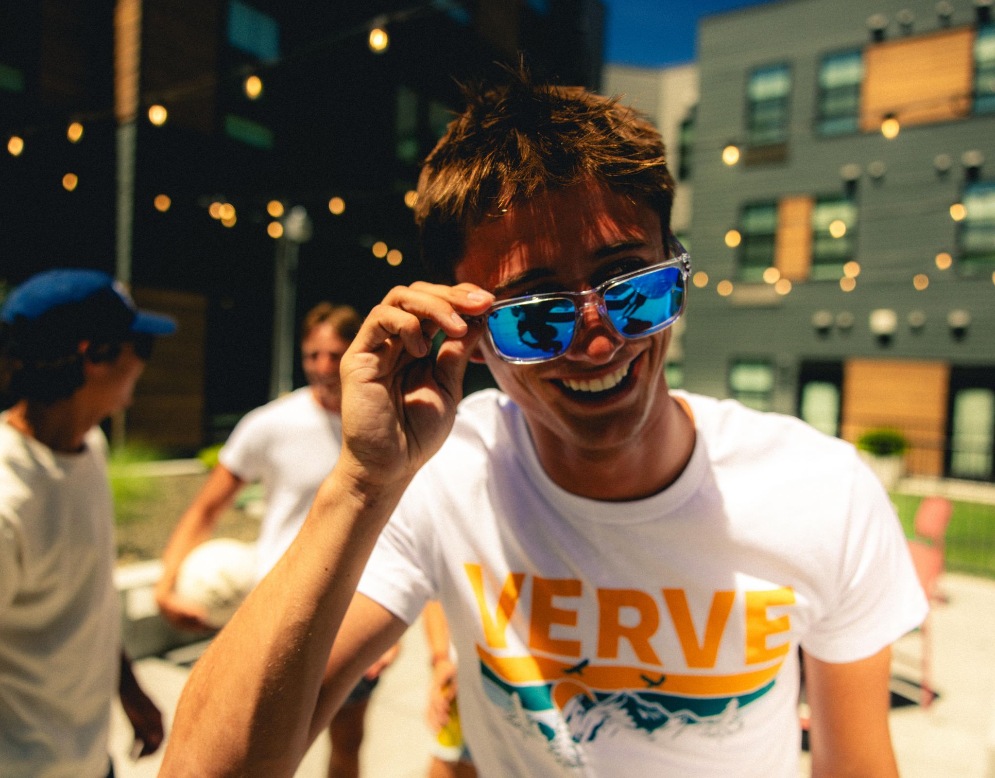 Smiling person in sunglasses and a "VERVE" shirt at an outdoor gathering with friends and string lights.
