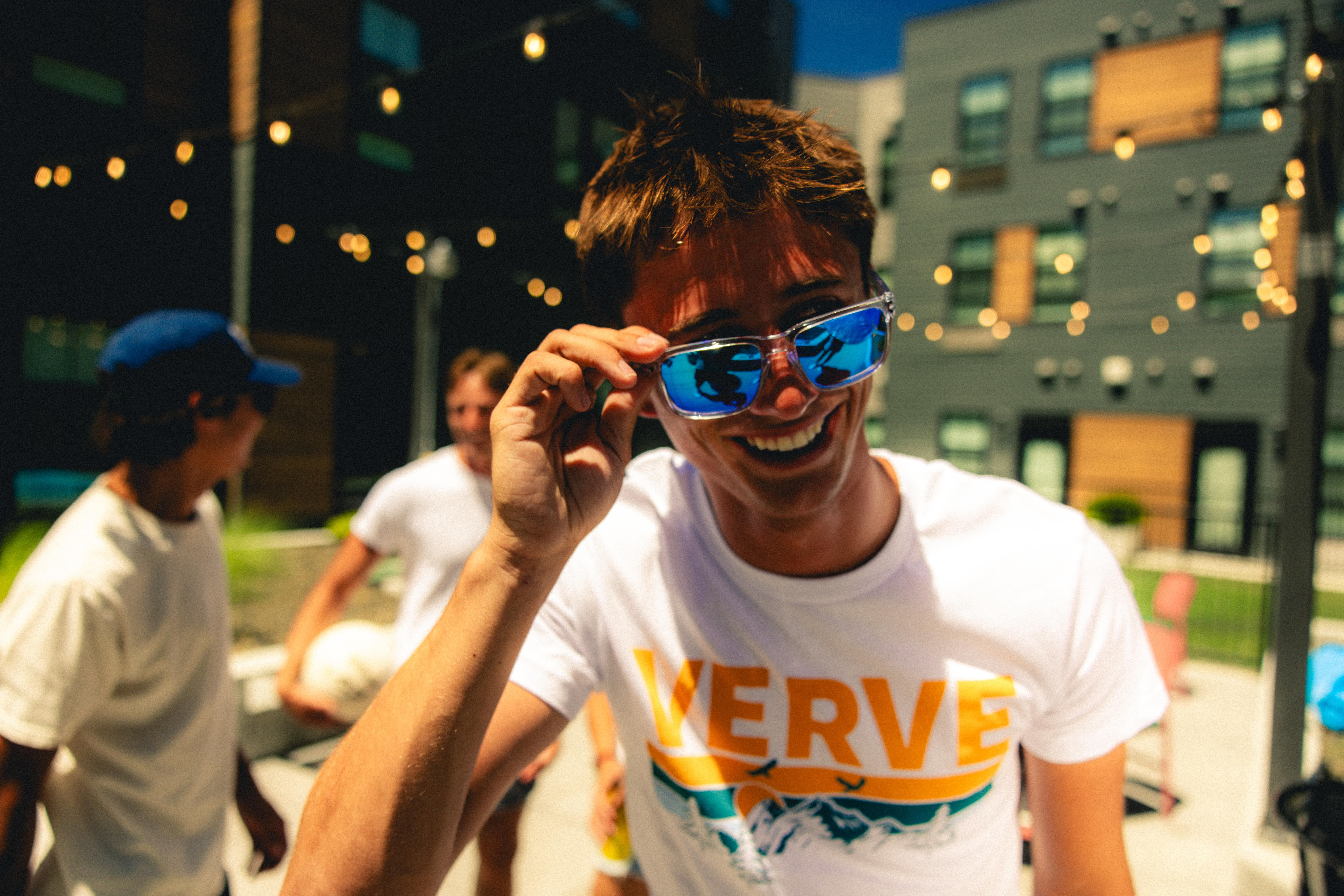 Smiling person in sunglasses and a "VERVE" shirt at an outdoor gathering with friends and string lights.