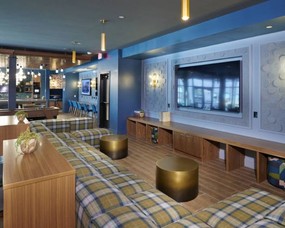 A spacious lounge at VERVE Boise featuring plaid couches, a large flat-screen TV, and a communal table with golden accent tables.